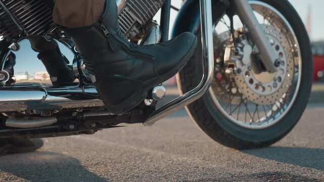 Close up of motorcyclist foot in black riding boot resting on bike stand, shiny chrome engine and front wheel brake disc visible, with blurred urban background of parked cars and buildings