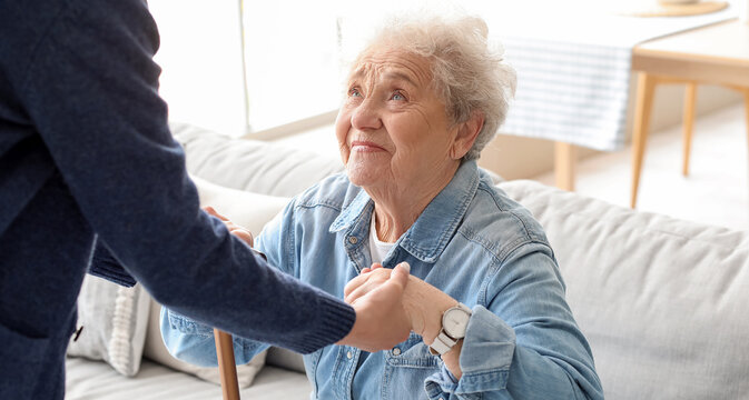 Young caregiver helping senior woman with stick to stand up at home