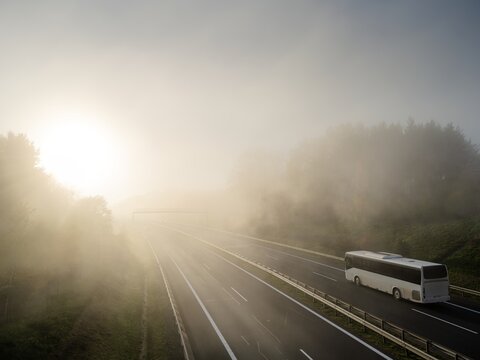 White bus driving on a highway in a forest shrouded in dense fog at sunset