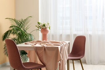 Vase with blooming branches on table near beige wall in dining room