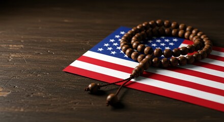On a dark wooden surface, string of brown prayer beads rests gently upon small American flag, illuminated by soft, somber light.