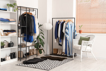 Fototapeta premium Armchair, shelving unit and racks with hanging clothes near white wall in room