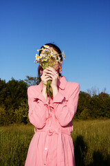 Young woman in a pink dress holding daisies in front of her face, standing in a sunny field,...