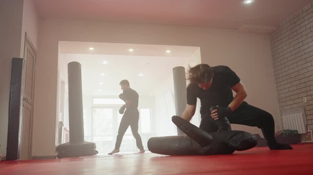 Athlete training in martial arts gym, one man practicing ground combat drills on dummy while another in background shadowboxes with gloves, showcasing power, stamina, discipline, concentration, and