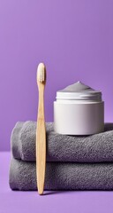 A bamboo toothbrush stands beside a jar of grey cream on a stack of grey towels against a lavender background