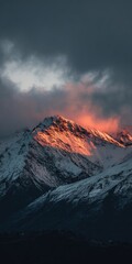 A snow-capped mountain peak glows with warm sunset hues under a dramatic, dark sky; the lower slopes are shadowed and cold