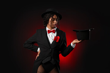 Young African-American magician showing magic trick with hat on dark red background
