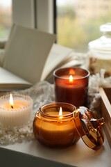 Burning candles, book and plaid on windowsill on rainy day, closeup. Autumn atmosphere