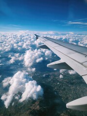 Airplane wing extending over a fluffy expanse of white clouds and a patchwork of green and brown terrain beneath a vibrant blue sky
