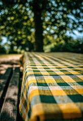 Close-up of a yellow and green checkered tablecloth on a picnic table outdoors, under a blurred leafy tree