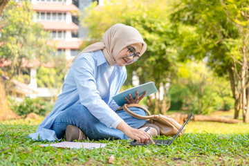 A young Asian Muslim woman sits on the bright green grass in a university park. She is diligently taking notes or journaling in a notebook, with a pen in hand, looking up with a warm, friendly smile.