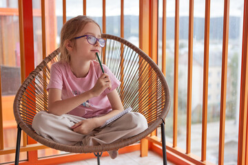 Girl with purple glasses sits in a modern chair, holding a pencil and notebook.
Thoughtful child gazes outside, inspired by imagination and curiosity.
Cozy indoor scene symbolizing creativity