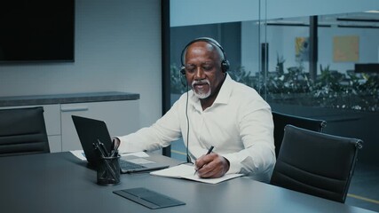 A mature African American businessman in a suit works diligently in a sleek corporate office. He takes notes while engaged in a call, surrounded by minimalistic decor and greenery. - Powered by Adobe