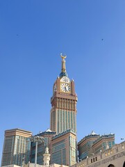 The impressive Makkah Royal Clock Tower and its adjacent minarets, featuring a golden crescent, against a vivid blue sky in Mecca, Saudi Arabia, during daylight.