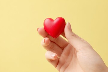 Fototapeta premium A delicate red heart held gently in a hand against a soft yellow backdrop.