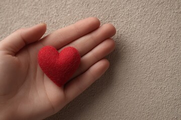 Fototapeta premium A small, textured, red felt heart rests gently in a light-skinned hand against a neutral beige textured surface.