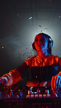 Slow motion vertical video of Asian female DJ in yellow jacket celebrating music moment, dancing and spinning turntable with golden confetti flying in smoky night club atmosphere