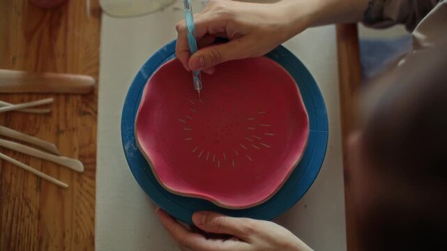 Hands of female potter using scribe tool to draw pattern on red glazed ceramic bowl placed on turntable in workshop. Top down view