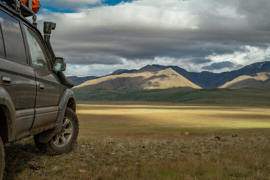 An off-road vehicle with mud tires, standing on a yellow field during the golden hour, with a view of a mountain and a cloudy sky. A travel vehicle. - Powered by Adobe
