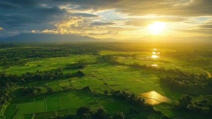 Obraz premium Aerial view of lush green farmland under a dramatic cloudy sky with the sun setting over distant mountains, casting golden light on the fields and creating a peaceful atmosphere
