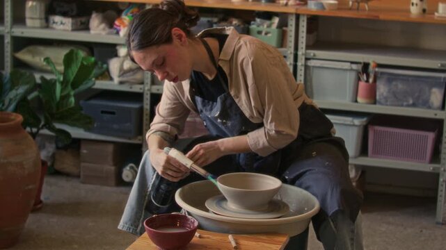 Young female ceramic artist sitting at pottery wheel, using handheld torch to dry clay bowl in cozy workshop