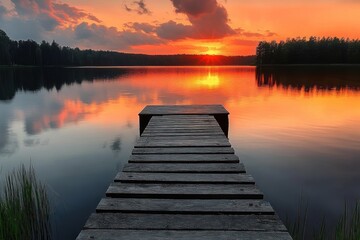 Fototapeta premium Wooden dock extending into calm lake with vibrant orange sunset and forest silhouette reflecting on water