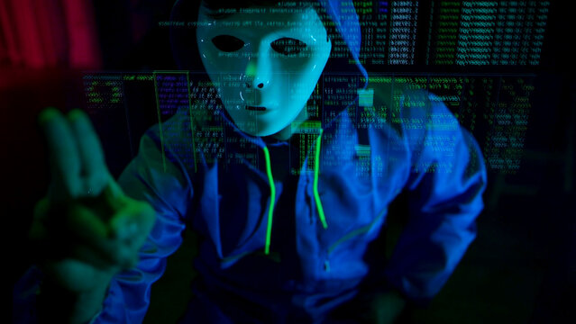 Anonymous hacker wearing a mask and hoodie interacting with digital data in a dark room