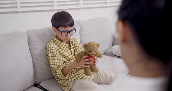 Asian boy with ADHD plays slowly with teddy bear on sofa during slowmotion therapy session in clinic while female psychologist observes, showing signs of emotional detachment and behavioral focus