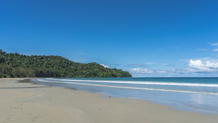 A beautiful tropical beach without people. The waves of the turquoise ocean foam and spread over the white sand. A green hill against a blue sky and clouds. Copy space. Malaysia. Borneo. Kota Kinabalu