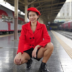 Asian woman in a red beret and coat crouches confidently on a train station platform, styled in black shorts and combat boots with urban trains in the background