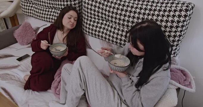 Two young women enjoy a relaxing morning at home, eating breakfast and watching television together on the sofa.