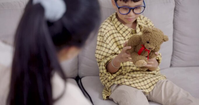 Asian boy with ADHD fidgets with teddy bear while seated on sofa in therapy session, engaging in behavioral self-soothing during psychological consultation with female therapist in clinic