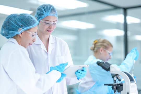 A senior scientist mentors a junior researcher, pointing at results on a digital tablet as they collaborate on an experiment in a bustling, modern scientific laboratory environment. - Powered by Adobe