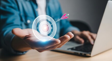 Person holding a target with arrow in bullseye on hand with laptop in background goal success