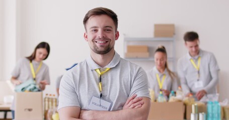 Portrait of a young happy smiling man volunteer looking cheerful at camera standing at charity center with people packing food grocery in donations box in background. 4k video. Slow motion video. - Powered by Adobe