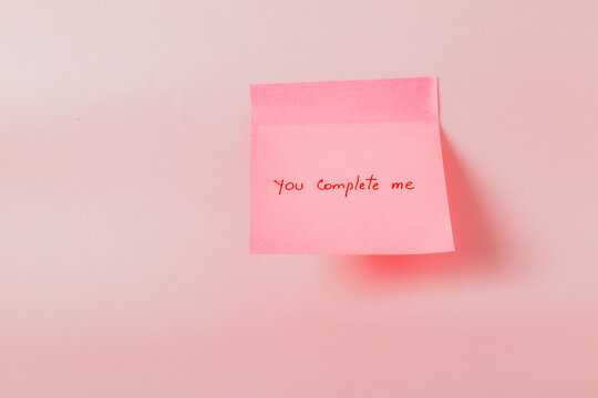 A pink sticky note with the words "You complete me" written on it. The note is hanging from a wall or a surface.