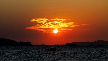 round sun sets over the sea, painting the sky in intense orange and deep red hues. Several boats...