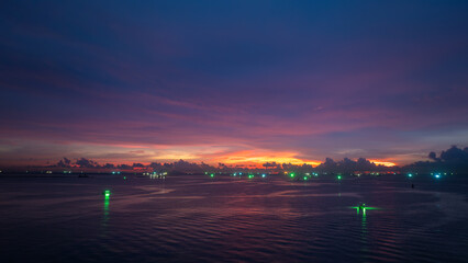 twilight time over the sea, The sunset sky is filled with deep purple, orange, and pink hues. Bright green lights from fishing and other boats twinkle on the water,