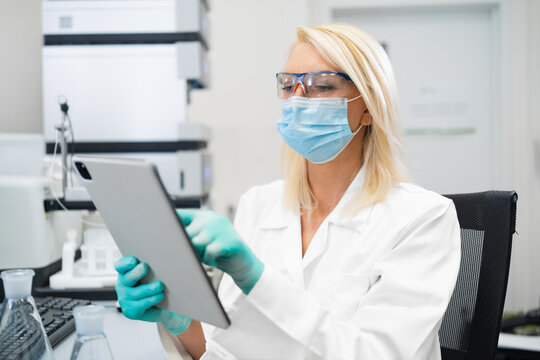 Female scientist in lab coat and mask using tablet to analyze data in laboratory.