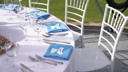 Elegant christening table setup with blue napkins, personalized tags, white chairs, and glassware arranged for a formal baby boy celebration in a bright, airy venue.