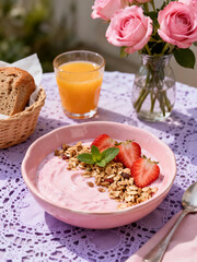A delightful outdoor breakfast featuring a pink bowl of yogurt with granola, strawberries, a glass of orange juice, and a vase of pink roses in the sun.