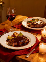 A dimly lit romantic dinner for two featuring juicy steaks with sauce, roasted vegetables, red wine, and candles for a cozy evening.