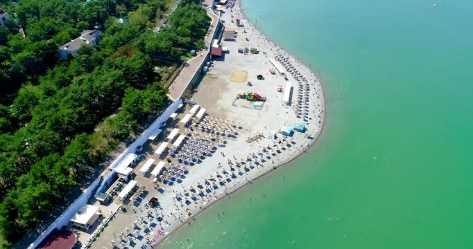 Gelendzhik Bay from bird's-eye view. Beach with umbrellas and sun loungers along sea. Embankment with balustrade. Yachts and ships at sea. Mountains in background. Sunset, long shadows. People swim an