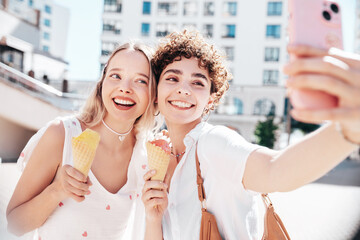 Two young beautiful smiling hipster female in trendy summer clothes. Carefree women posing on street background. Positive models eating tasty ice cream in waffles cone in sunny day, cheerful and happy