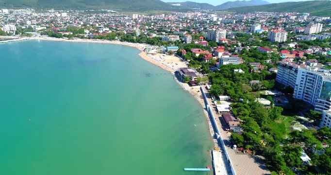 Flight over central beach resort of Gelendzhik. Promenade with balustrade runs along beach. Umbrellas and sunbeds on beach. Blue sea. Caucasus Mountains in background.