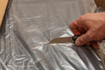 Close up of a mans hand using a knife to open a cardboard box sealed with clear tape.