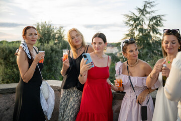 Group of women in elegant dresses drinking cocktails at sunset