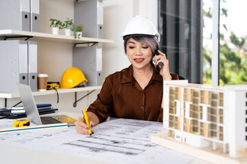 Senior foreman or civil engineer in safety helmet Talking on phone call working at a desk in construction site office with Construction blueprint drawings plans .