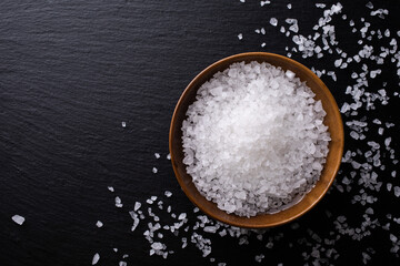 Pile of sea salt in ceramic bowl on black stone kitchen table top view.