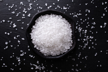 Pile of sea salt in ceramic bowl on black stone kitchen table top view.